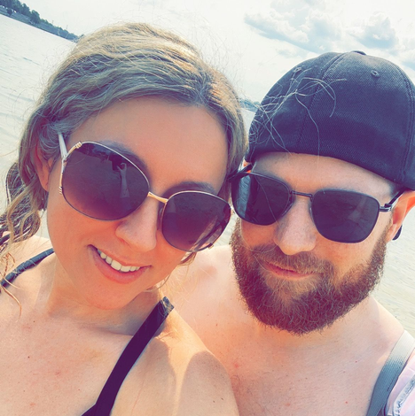 Couple in sunglasses smiling, near water on a sunny day. Woman's arm around man.