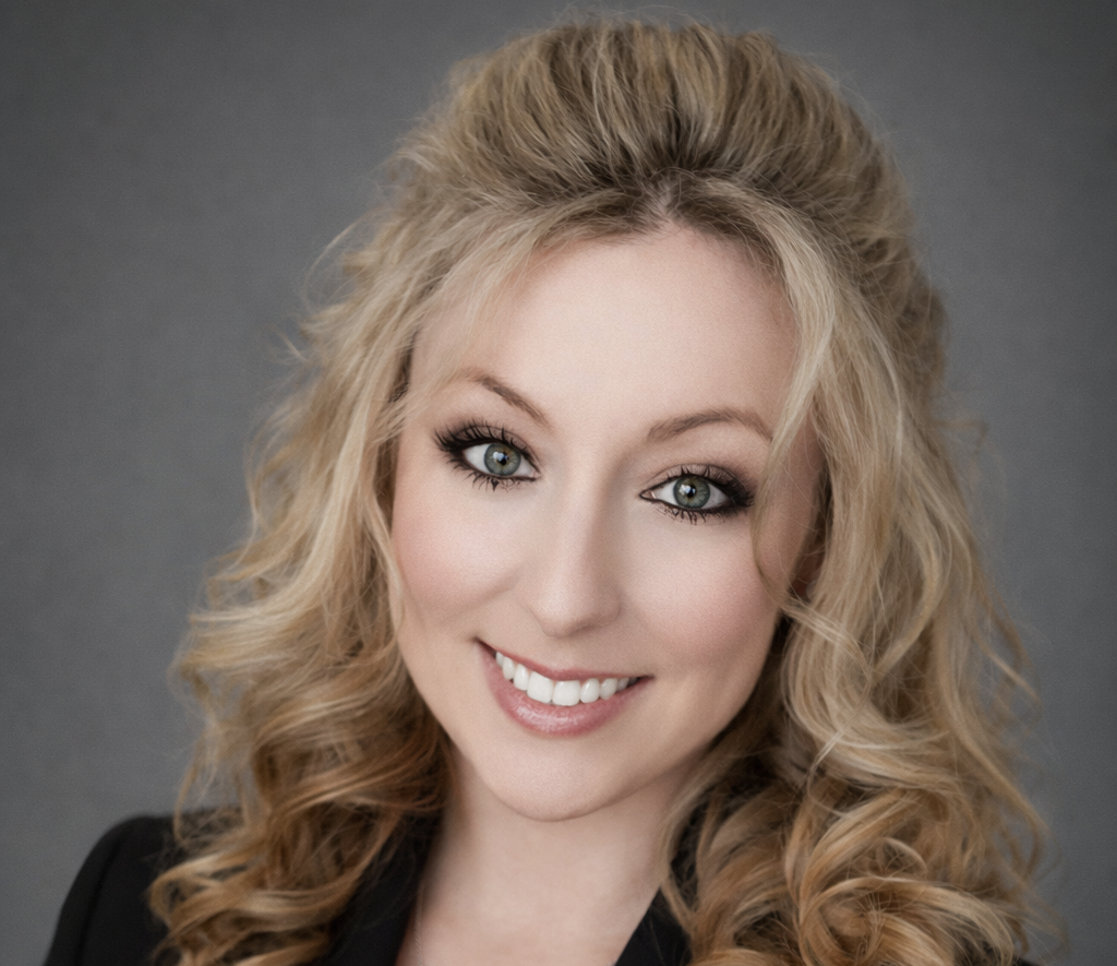 Woman with blonde curly hair, smiling with green eyes, wearing a dark jacket, against a grey background.