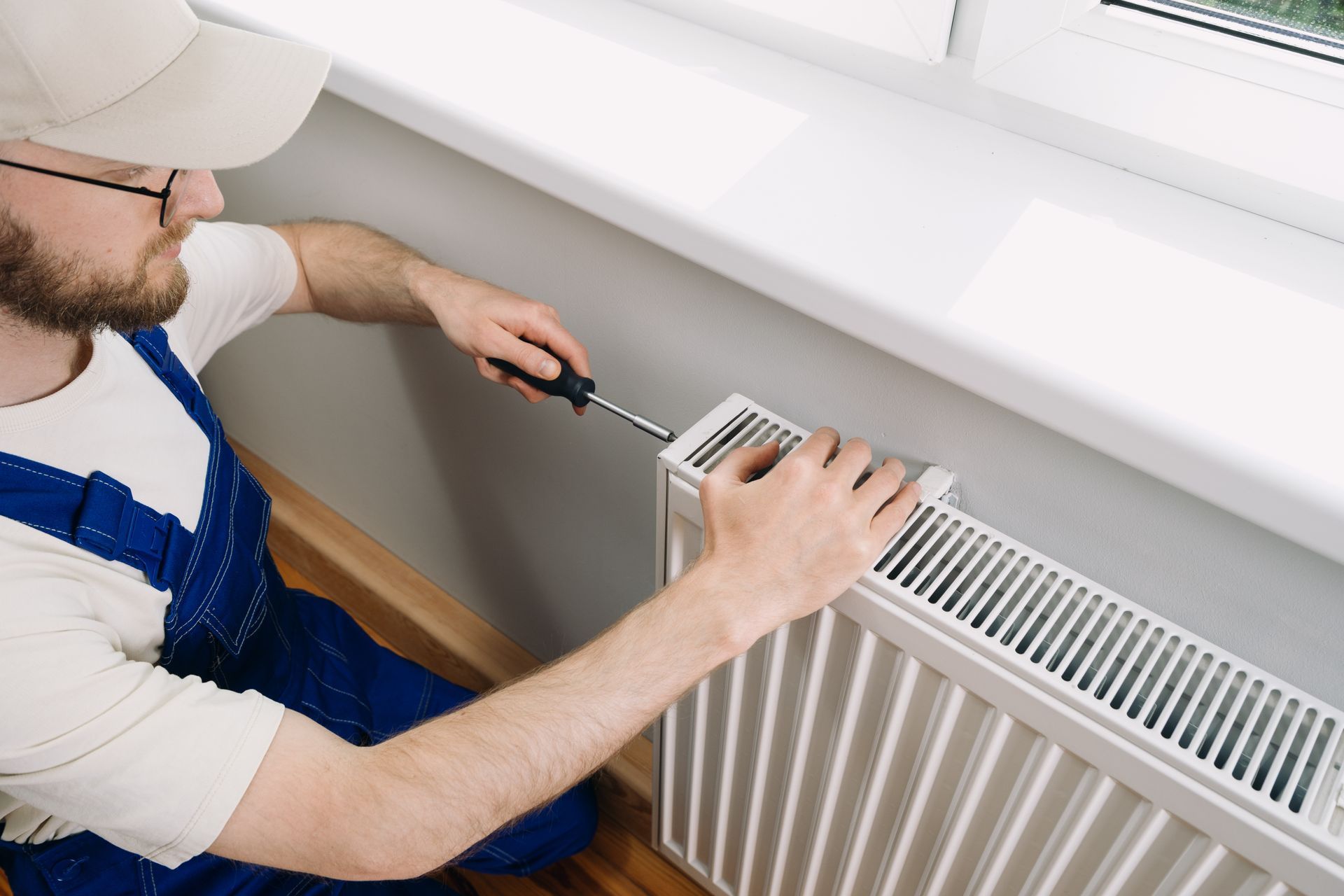 Technician performing heating system repair on white radiator near window with screwdriver.