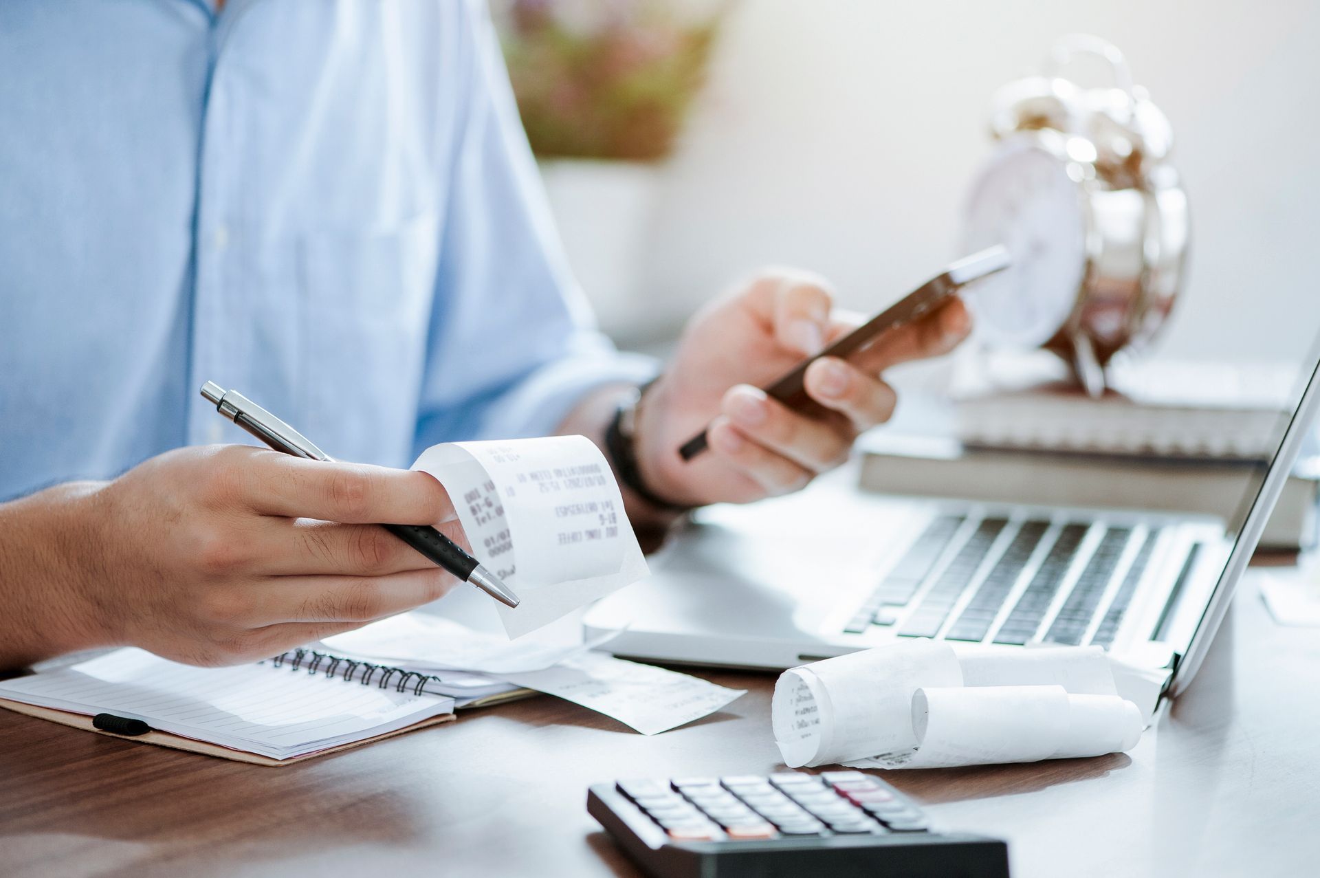 A man is sitting at a desk holding a receipt and using a cell phone.