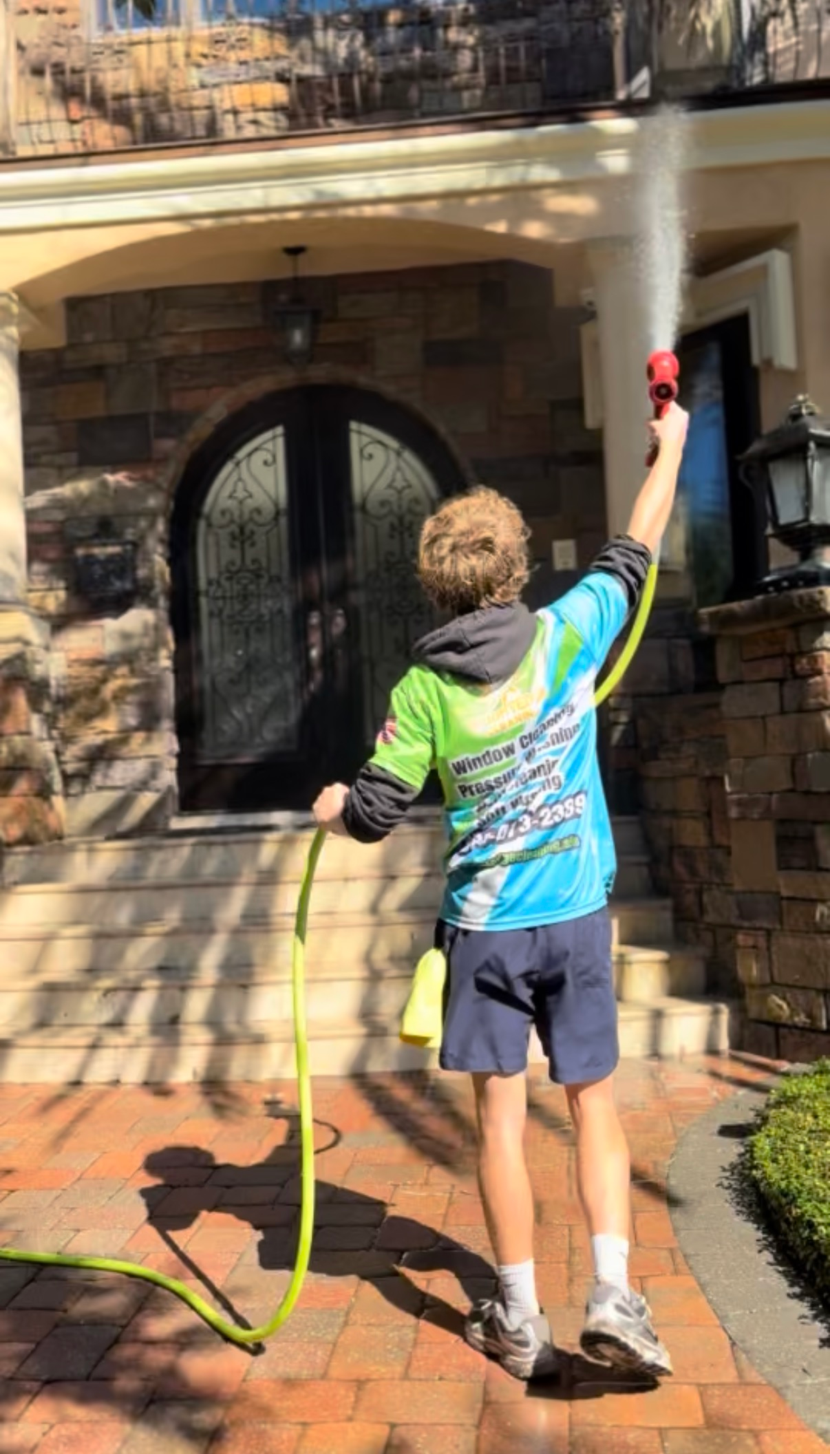 Person sprays water towards a house; wearing tie-dye shirt and shorts, holding hose.