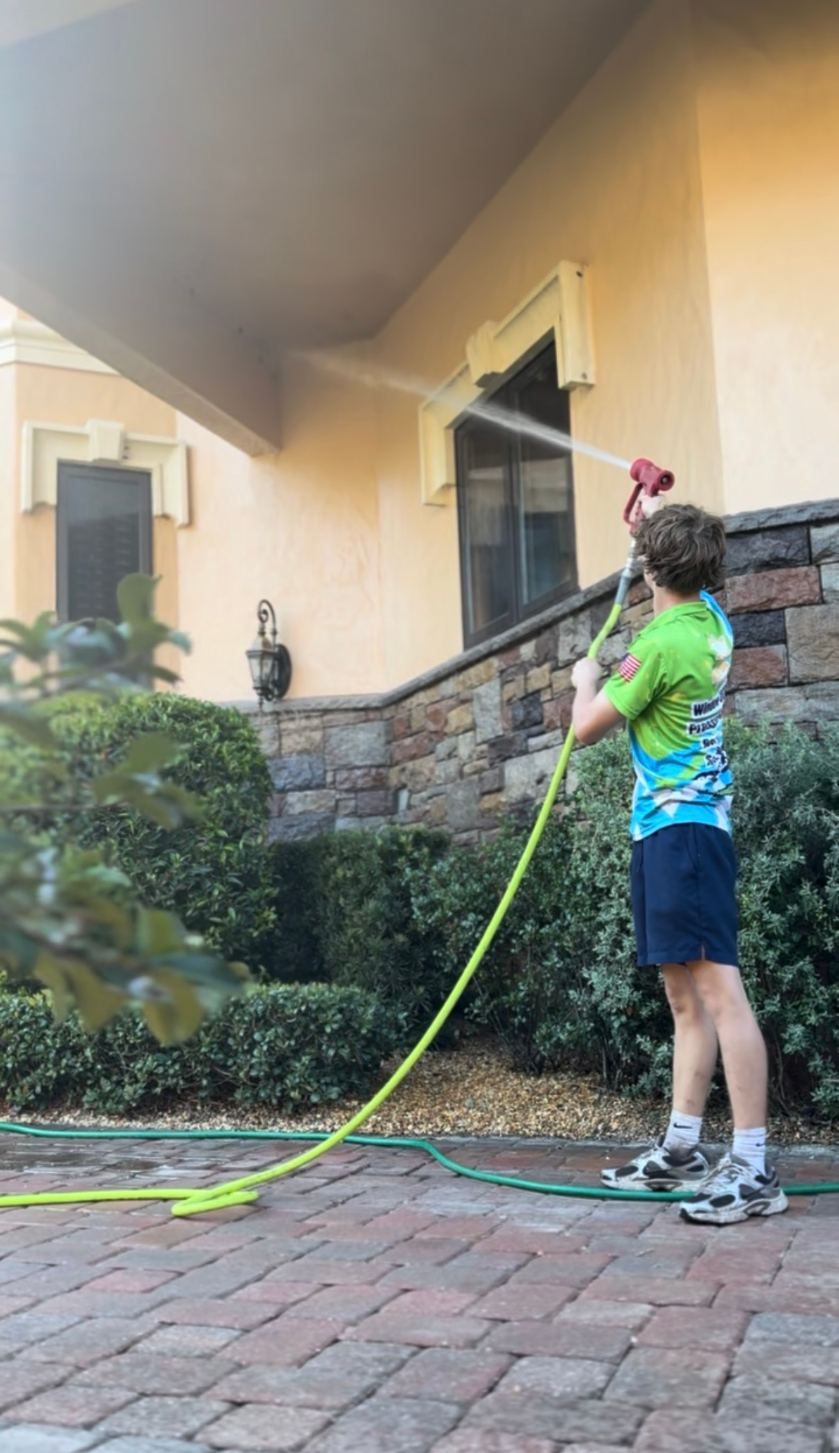 Boy spraying water on a building's exterior. The building is tan and has a stone wall base.