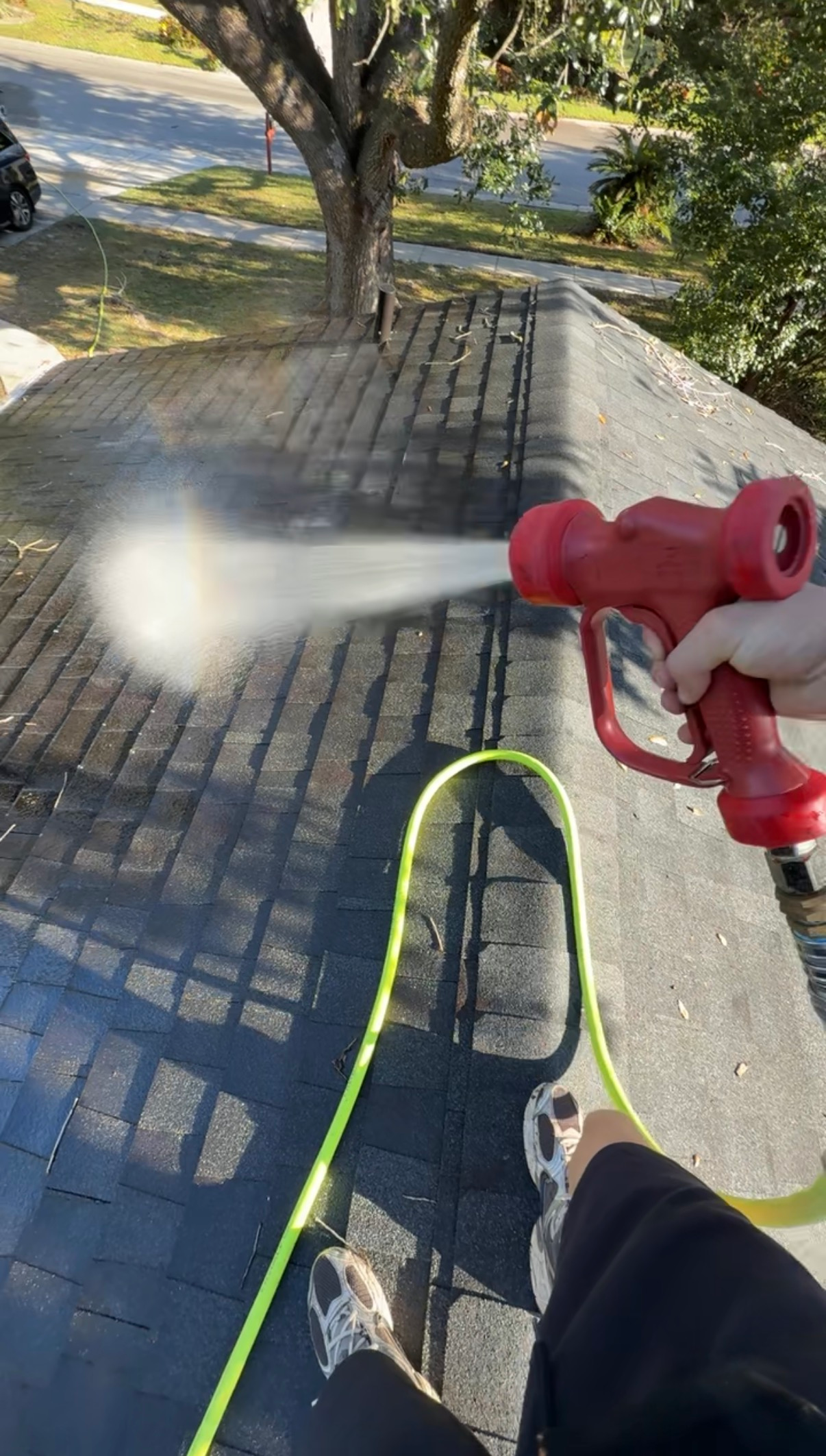 Person using a red pressure washer on a roof, spraying water. Green hose and black asphalt shingles.