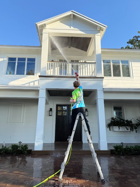 Person pressure washing a white house balcony from a ladder.