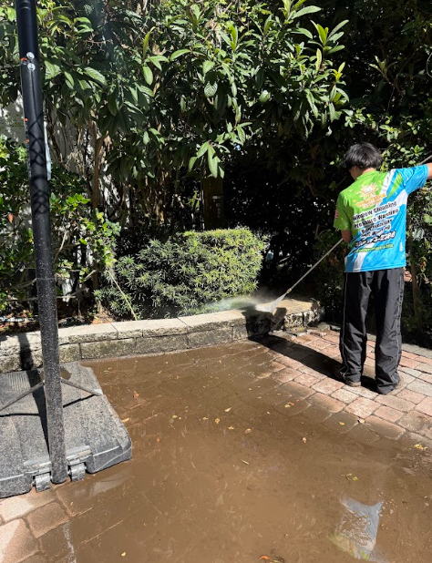 Person power washing a brick patio and a retaining wall outdoors on a sunny day.