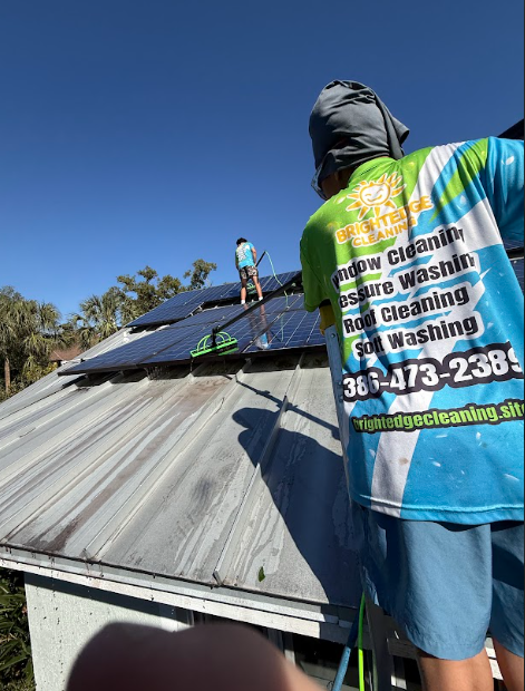 Two workers cleaning solar panels on a rooftop; one is spraying. Sunny day, blue sky.