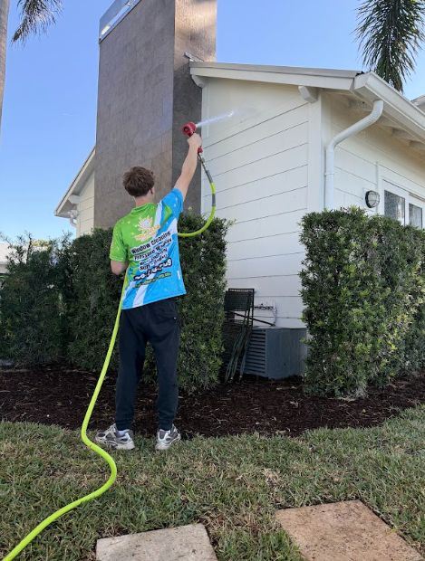 Person spraying side of house with a hose; green and white shirt, green hose, beige house, sunny outdoors.