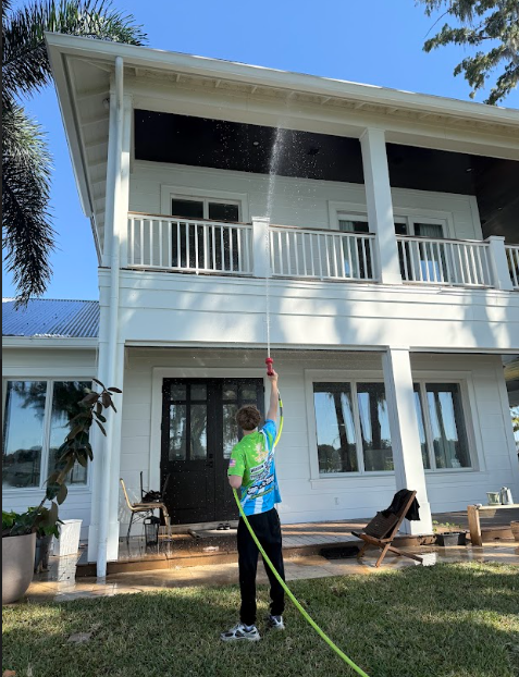 A person pressure washing a white two-story house with black trim on a sunny day.