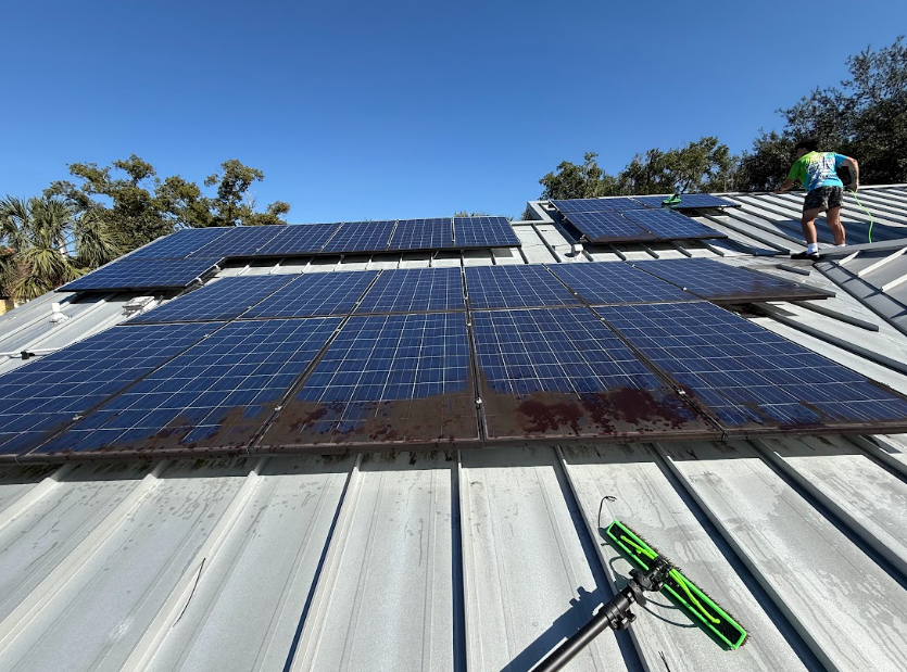 Person cleaning solar panels on a metal roof under a blue sky.