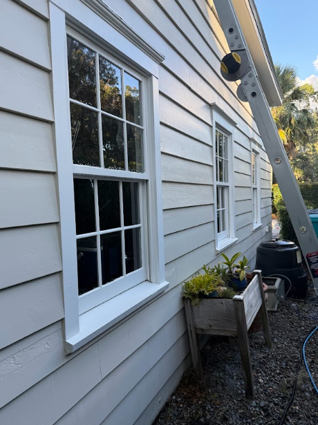 Exterior of a house with white windows and siding. A ladder leans against the building. A planter sits below.