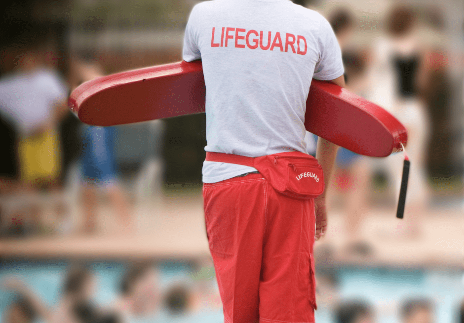 A lifeguard is standing in front of a pool holding a life preserver.