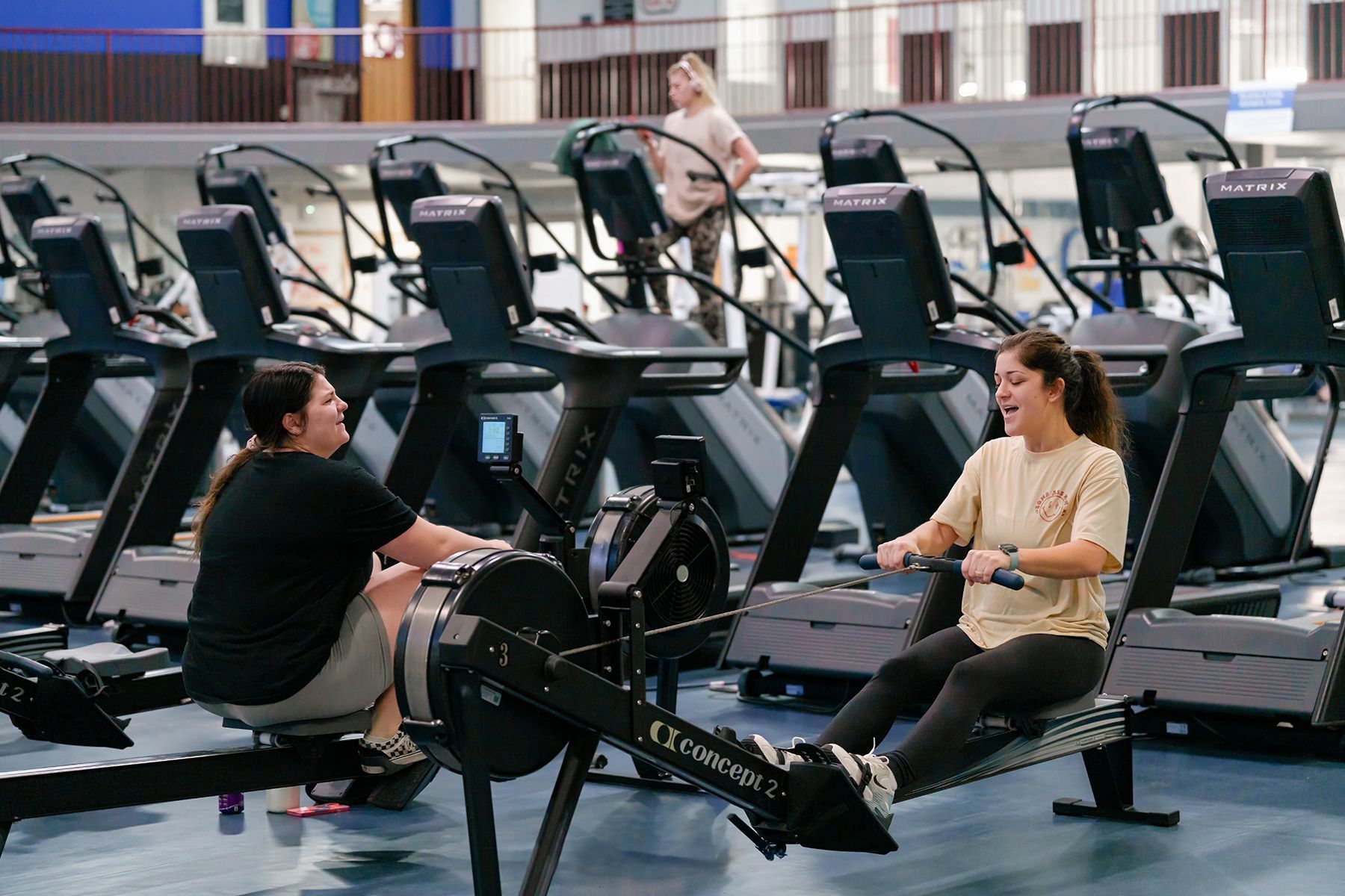 Two women are sitting on rowing machines in a gym.