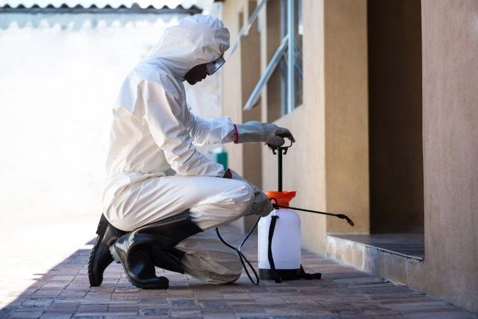 Person in white protective suit spraying a building wall with a pump sprayer outdoors