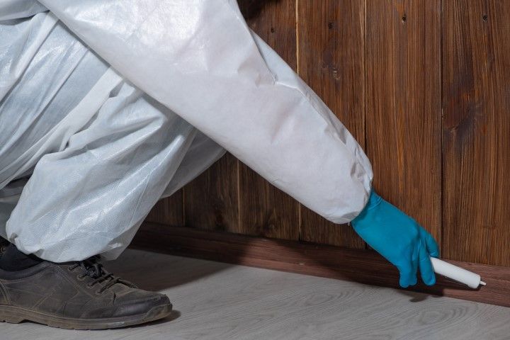 Gloved worker using a caulk gun along a wooden floor edge beside a wall