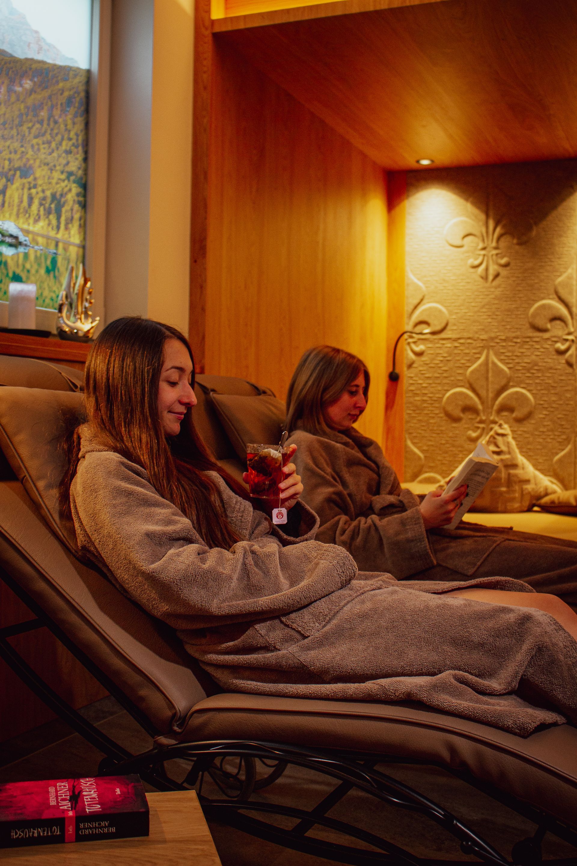 Two women are sitting in lounge chairs in a room reading books and drinking wine.