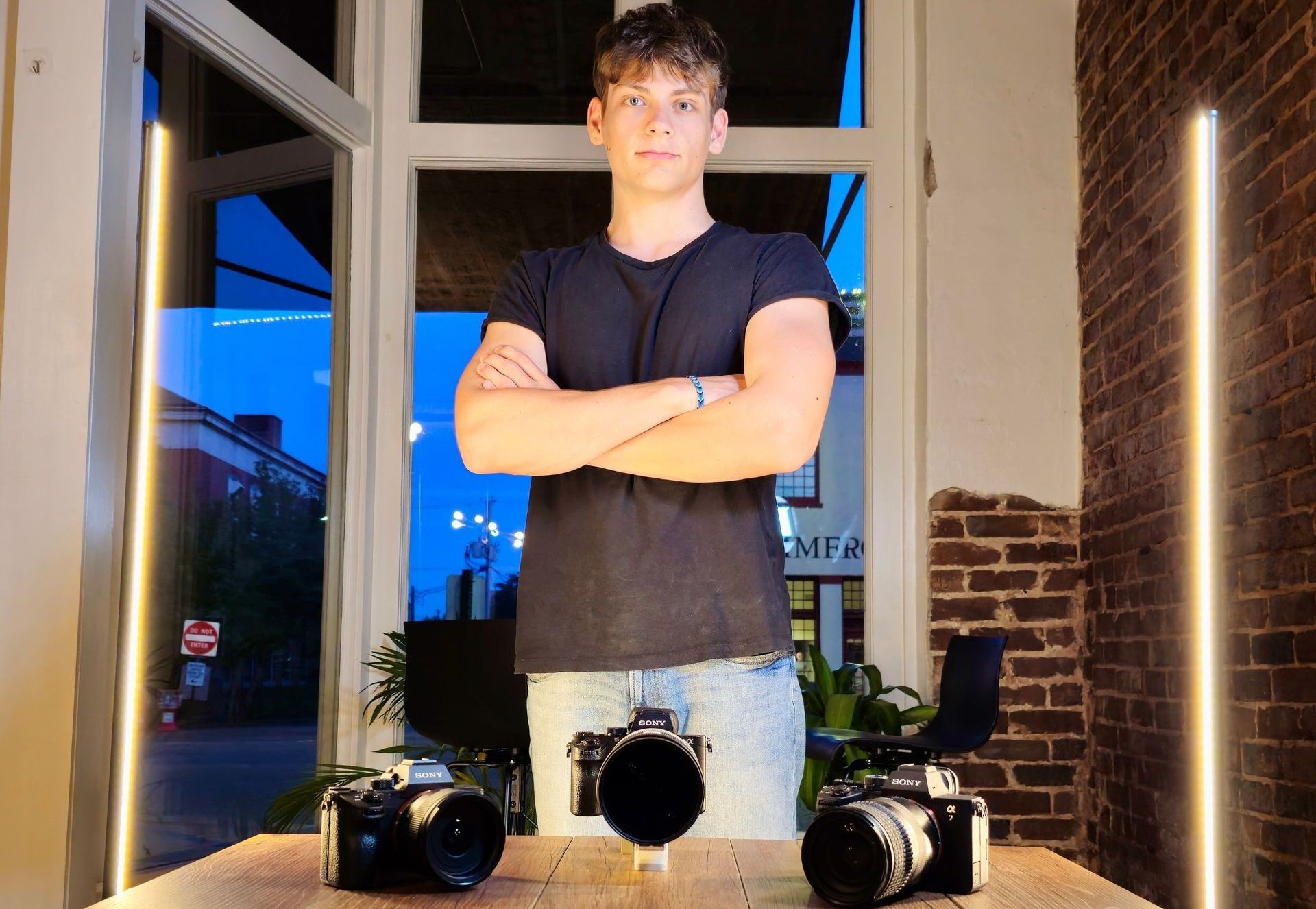 A man is standing with his arms crossed in front of two cameras on a table.
