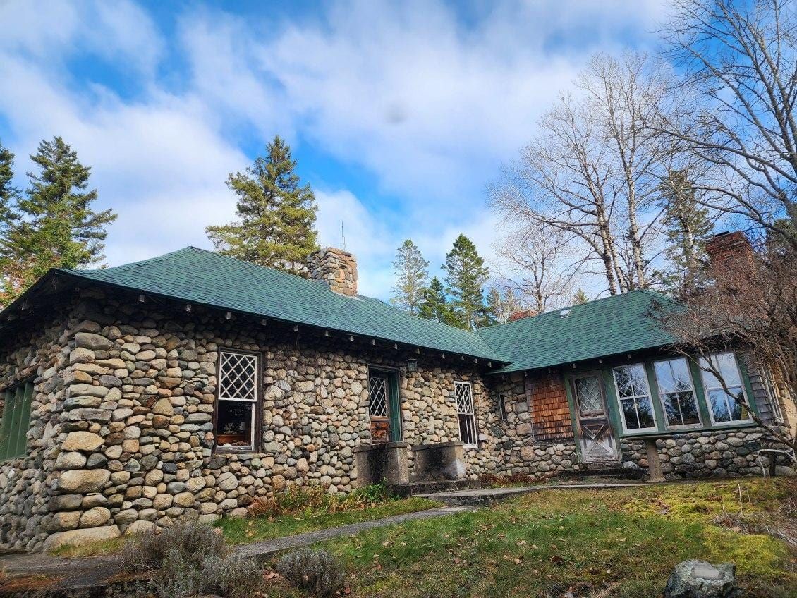 A large stone house with a green roof is surrounded by trees.