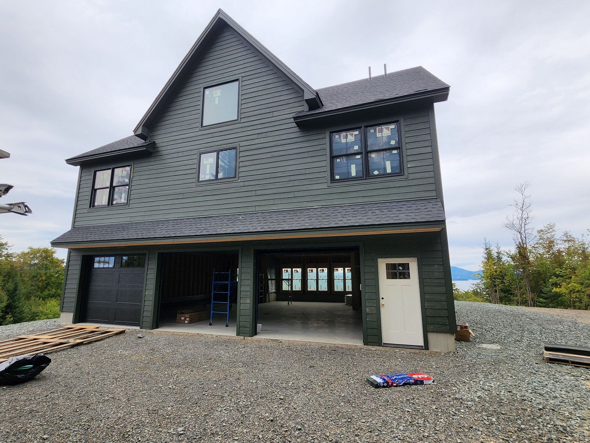 A multi-story house with dark gray shingles under construction, featuring an open garage and gravel driveway.