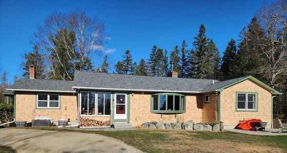 A large brick house with a gray roof is surrounded by trees on a sunny day.