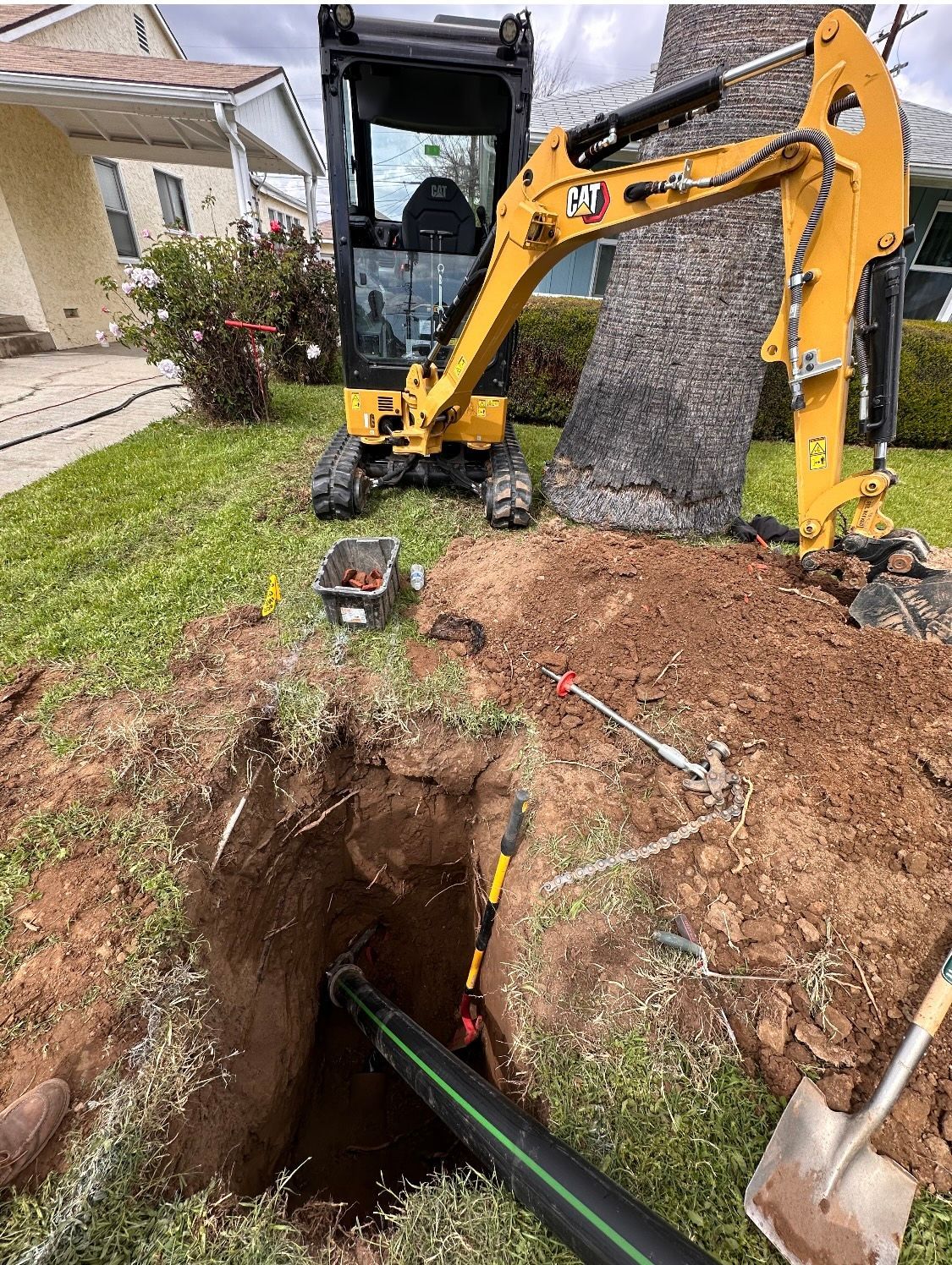 A yellow excavator is digging a hole in the ground in front of a house for sewer repair.