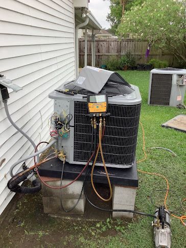 Air conditioner unit being serviced with gauges, hoses, and a vacuum pump, outdoors near a house.