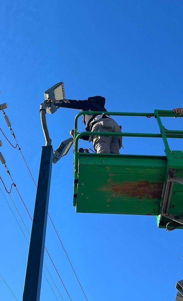 Person on a lift working on a light fixture attached to a tall pole, against a blue sky.