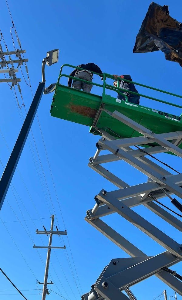 Workers on a green lift installing a street light against a blue sky, near power lines.