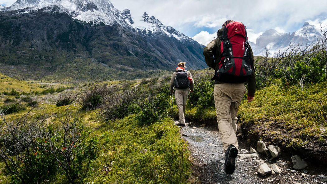 Two hikers with backpacks walk along a mountain trail toward snow-capped peaks.