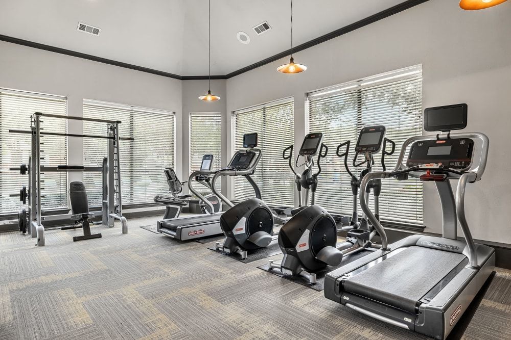 Interior of a modern gym with various exercise equipment, including treadmills, elliptical trainers, a stationary bike, and a weightlifting rack at Marquis at Stonebriar in Frisco, TX.