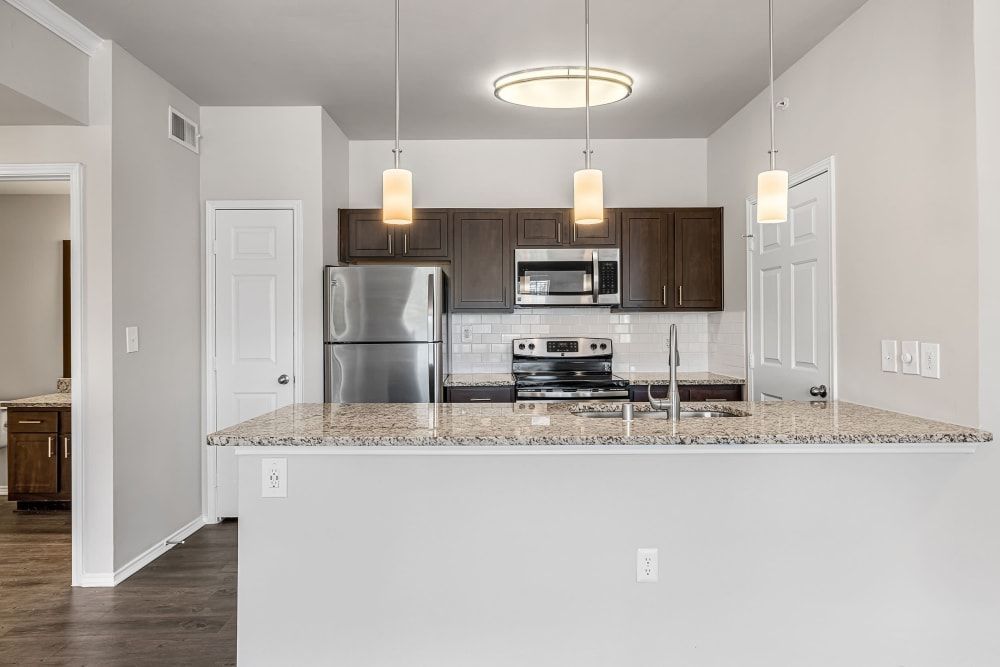 Modern kitchen interior with a granite countertop island, stainless steel refrigerator, stove, and microwave, dark wood cabinets, and three pendant lights hanging above the island. The room has light gray walls and dark flooring, with a view into an adjoining room at Marquis at Stonebriar in Frisco, TX.
