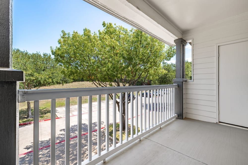 View from a shaded apartment balcony with a white railing, looking out onto a landscaped area with green trees and a parking lot on a sunny day at Marquis at Stonebriar in Frisco, TX.