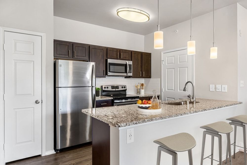 Modern kitchen interior with stainless steel appliances, dark wood cabinets, and granite countertops. There's a breakfast bar with stools, pendant lighting above, and a bowl of fruit and a jug on the counter at Marquis at Stonebriar in Frisco, TX.
