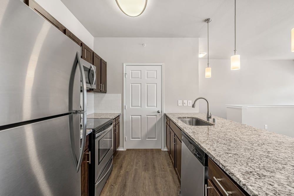A modern kitchen interior with stainless steel appliances, dark wood cabinets, granite countertops, and wooden flooring. The space is well-lit with a combination of ceiling lights and stylish pendant lights above the counter at Marquis at Stonebriar in Frisco, TX.