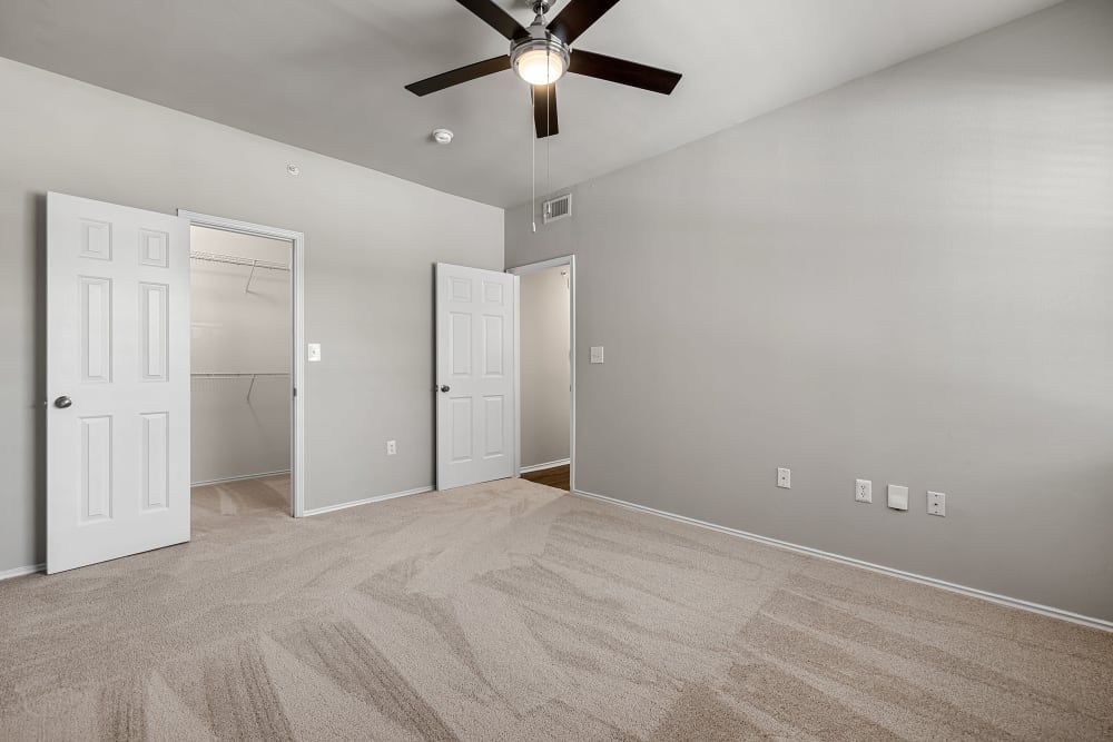 Empty modern room with beige carpet flooring, white walls, and a ceiling fan with lights. On the left, there's a closed white door and an open closet with built-in shelving. The right side has another closed white door and electrical outlets along the wall. There is no furniture or personal items, giving the space a clean, vacant look at Marquis at Stonebriar in Frisco, TX.