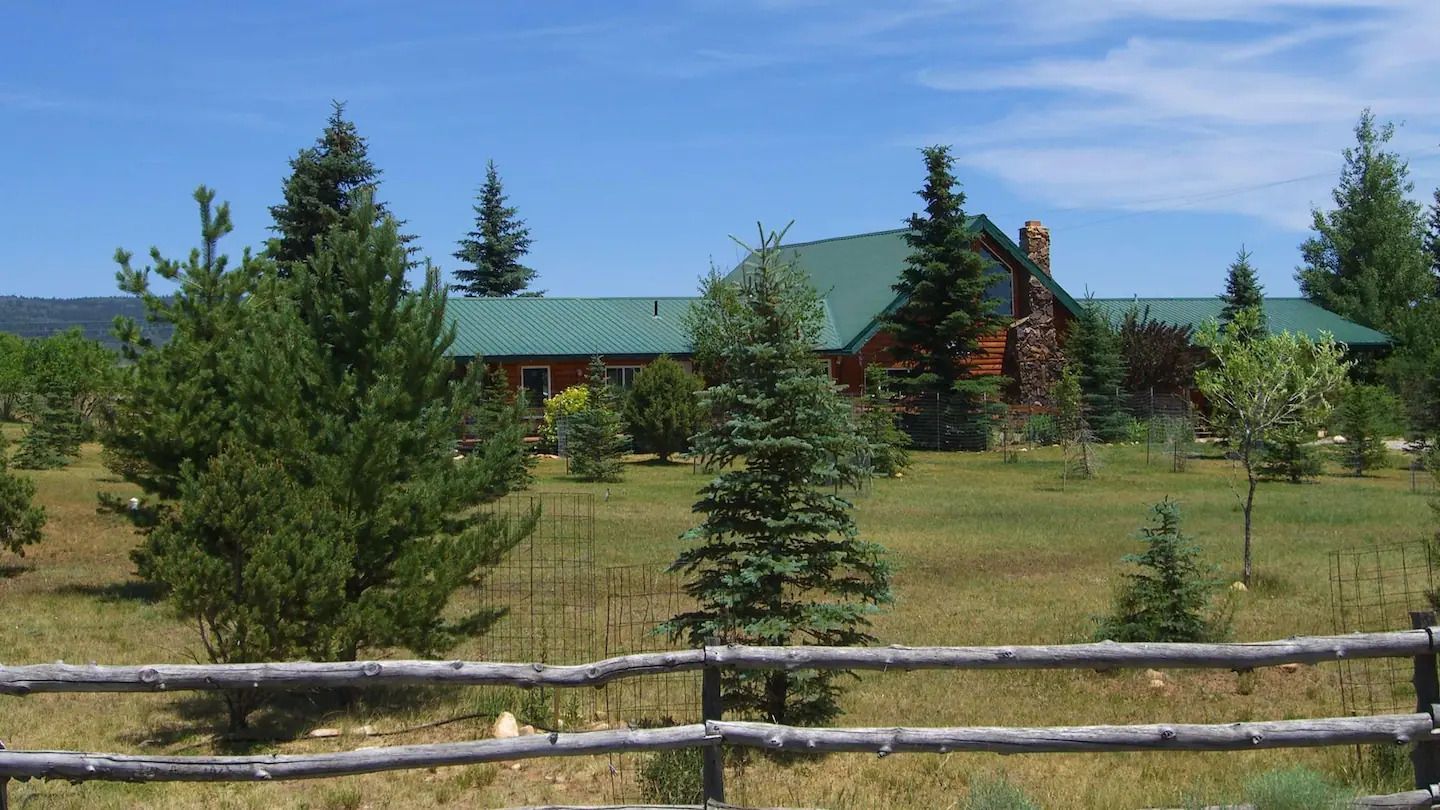 A house with a green roof is surrounded by trees and a wooden fence