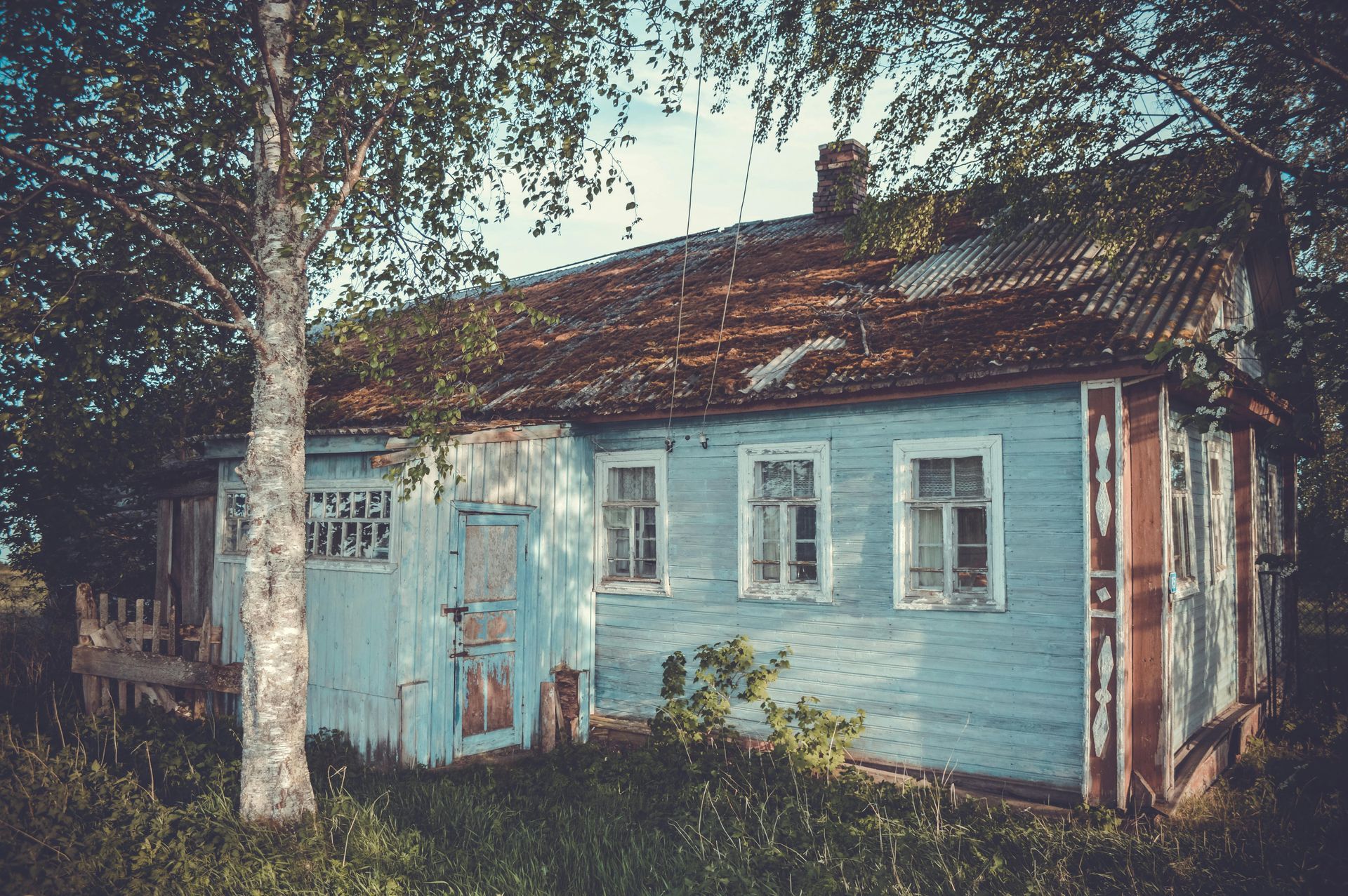 A weathered, light blue wooden house with a rusted roof, surrounded by trees and overgrown grass.