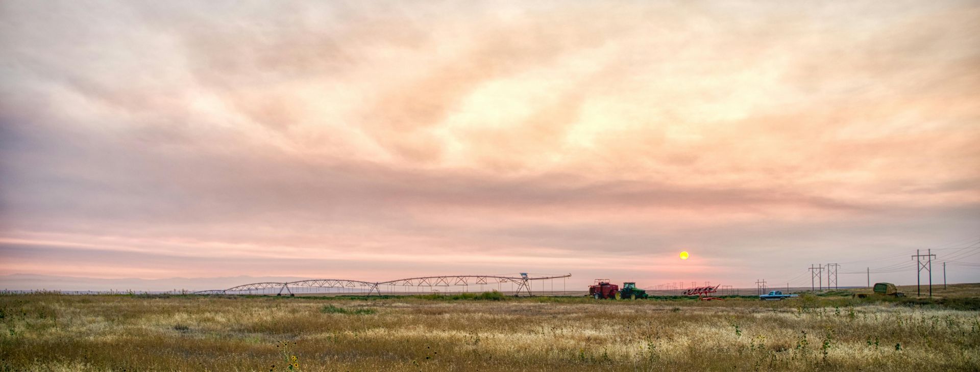 A sunrise over a flat, rural landscape with a farm irrigation system and a distant tractor under a hazy, pastel sky.