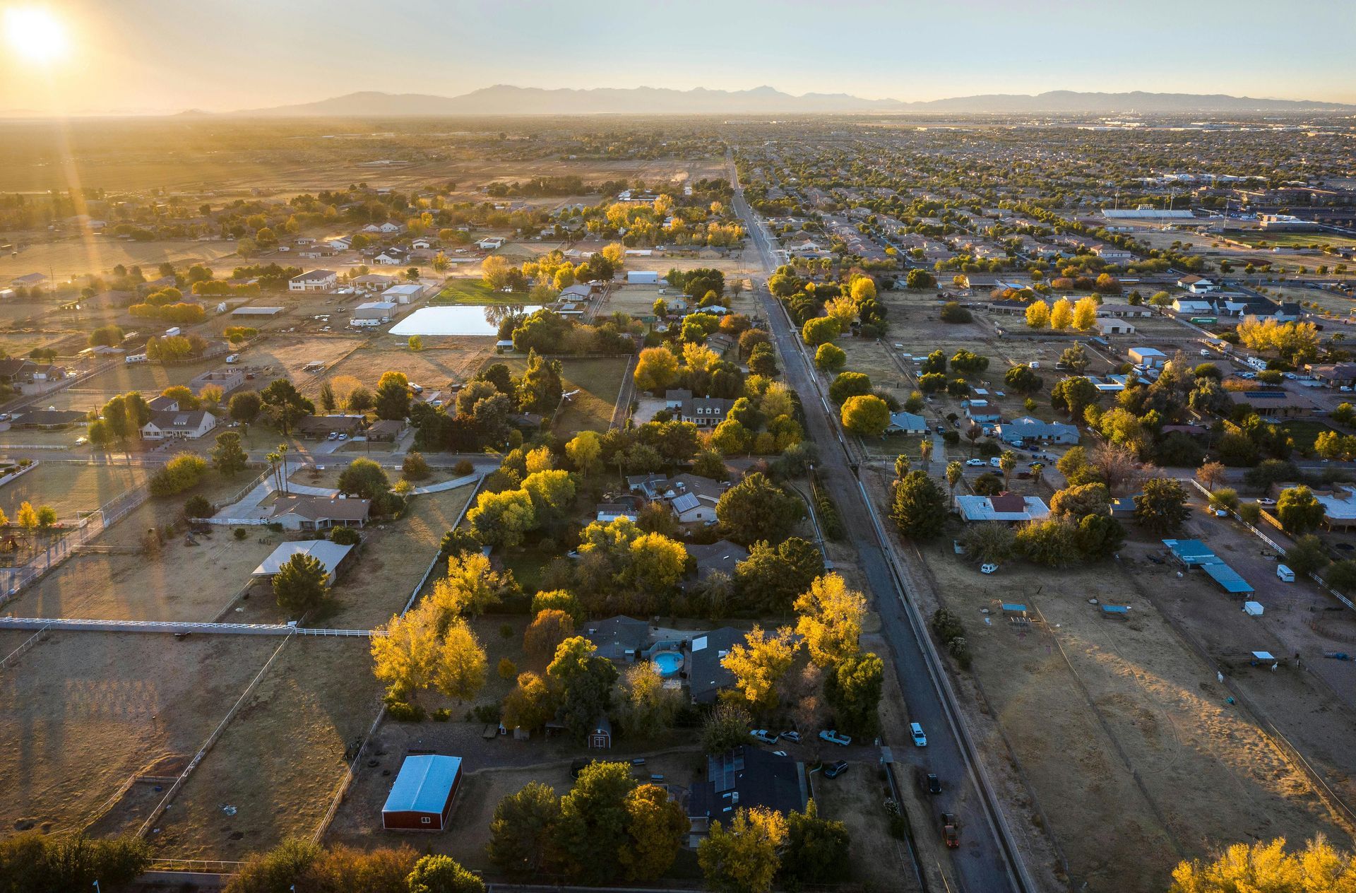 Aerial view of a residential neighborhood with golden trees at sunset, featuring a long road and mountains in distance.
