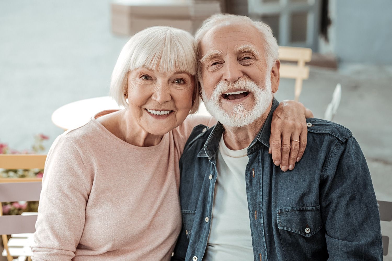 Elderly couple smiling and embracing outdoors. Woman in pink, man in denim shirt.