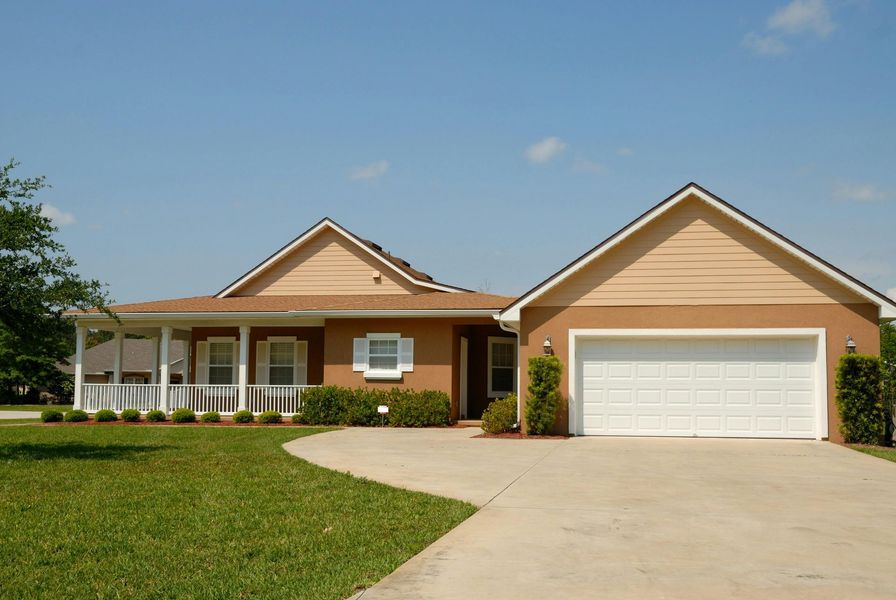 Tan stucco house with white porch and garage. Concrete driveway, green grass, blue sky.