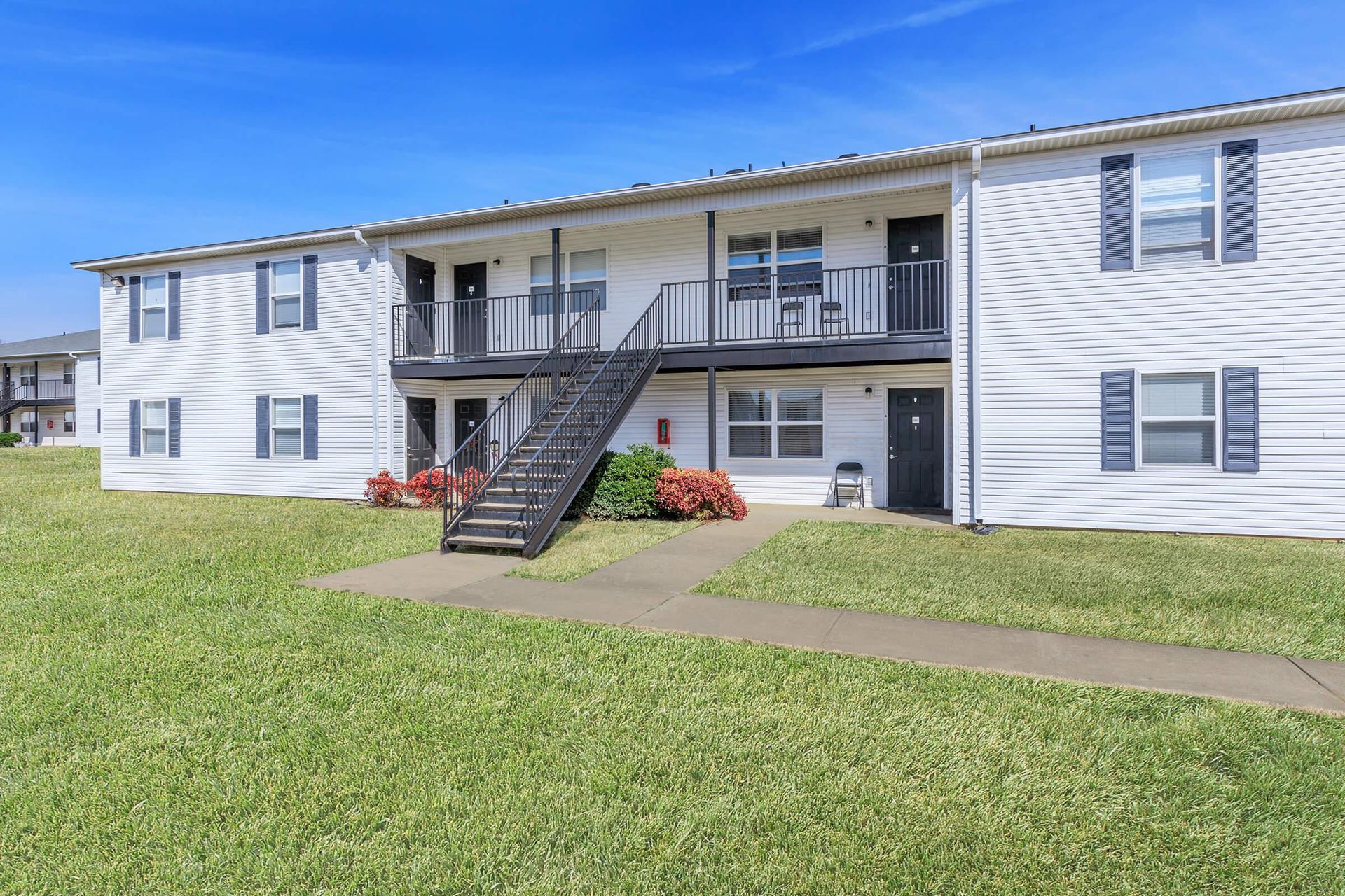 Two-story white apartment building with black stairwell, green lawn, and blue shutters against a blue sky.
