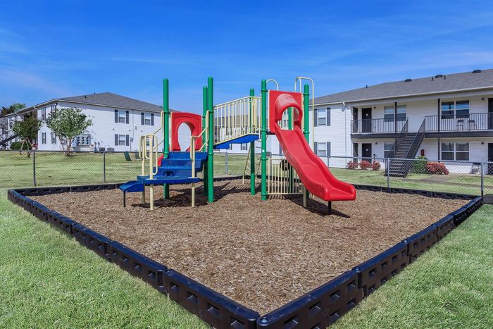 Playground with red slide, blue steps, and green supports on wood chips, surrounded by grass, and two-story apartments.