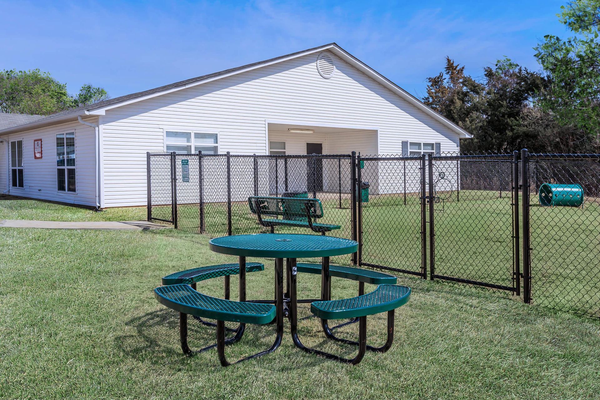 Green picnic table in a grassy dog park enclosed by a black fence in front of a white building.