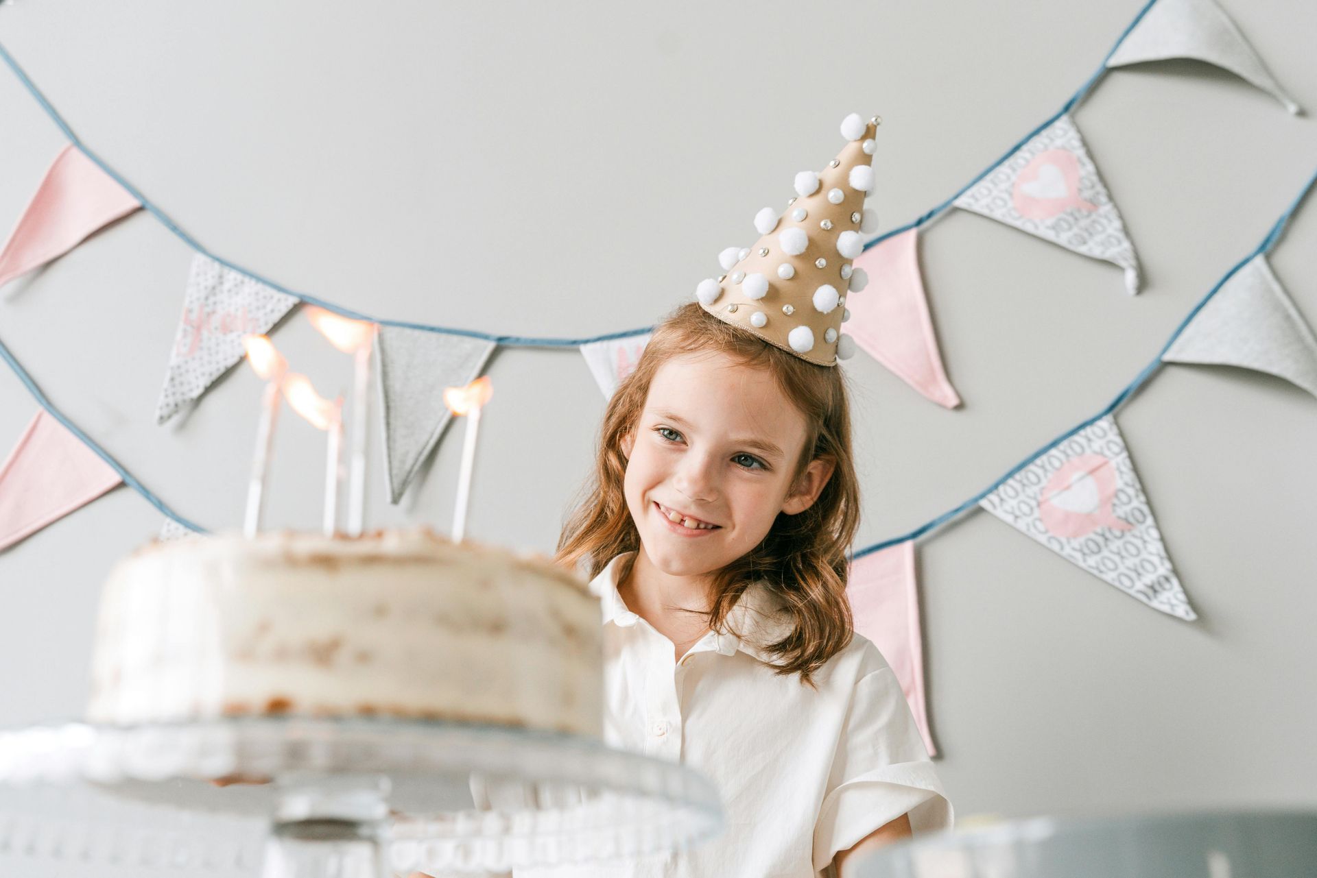 A little girl wearing a party hat is sitting in front of a birthday cake.