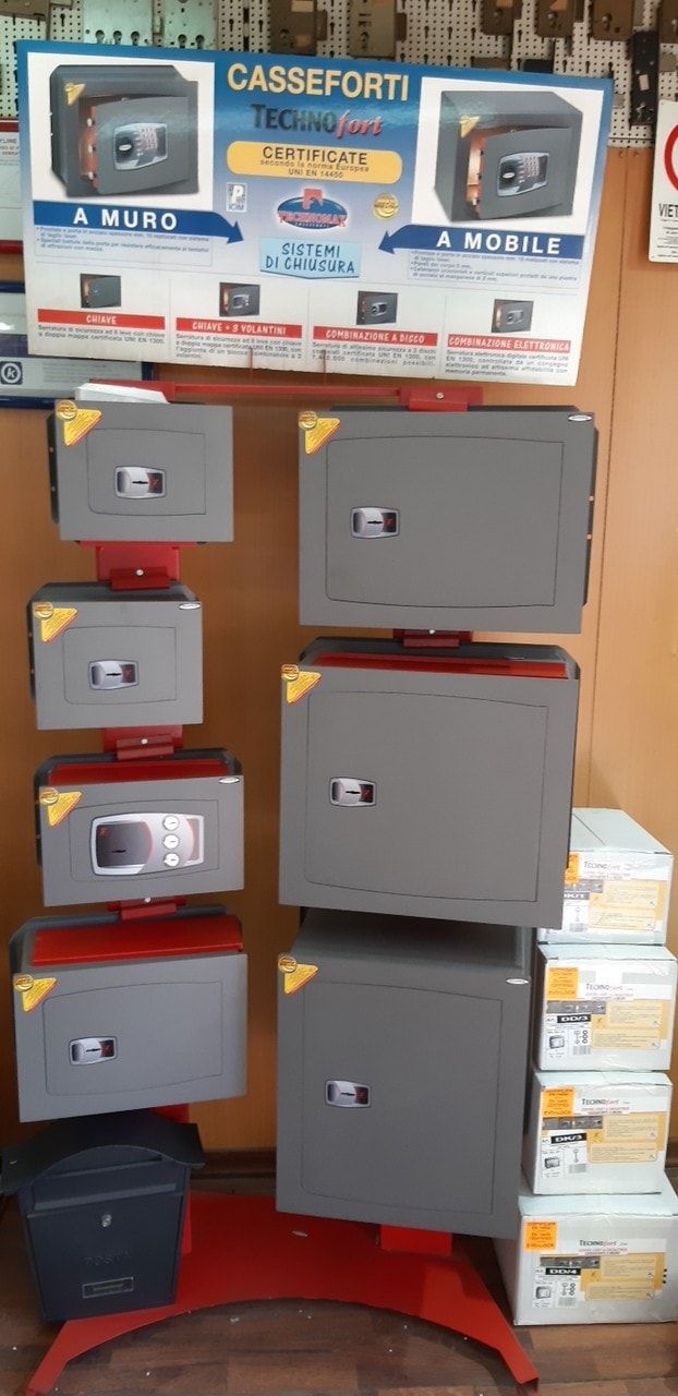 Esposizione di varie casseforti grigie, alcune impilate e altre su un supporto rosso a più livelli, con un cartello promozionale sopra.
