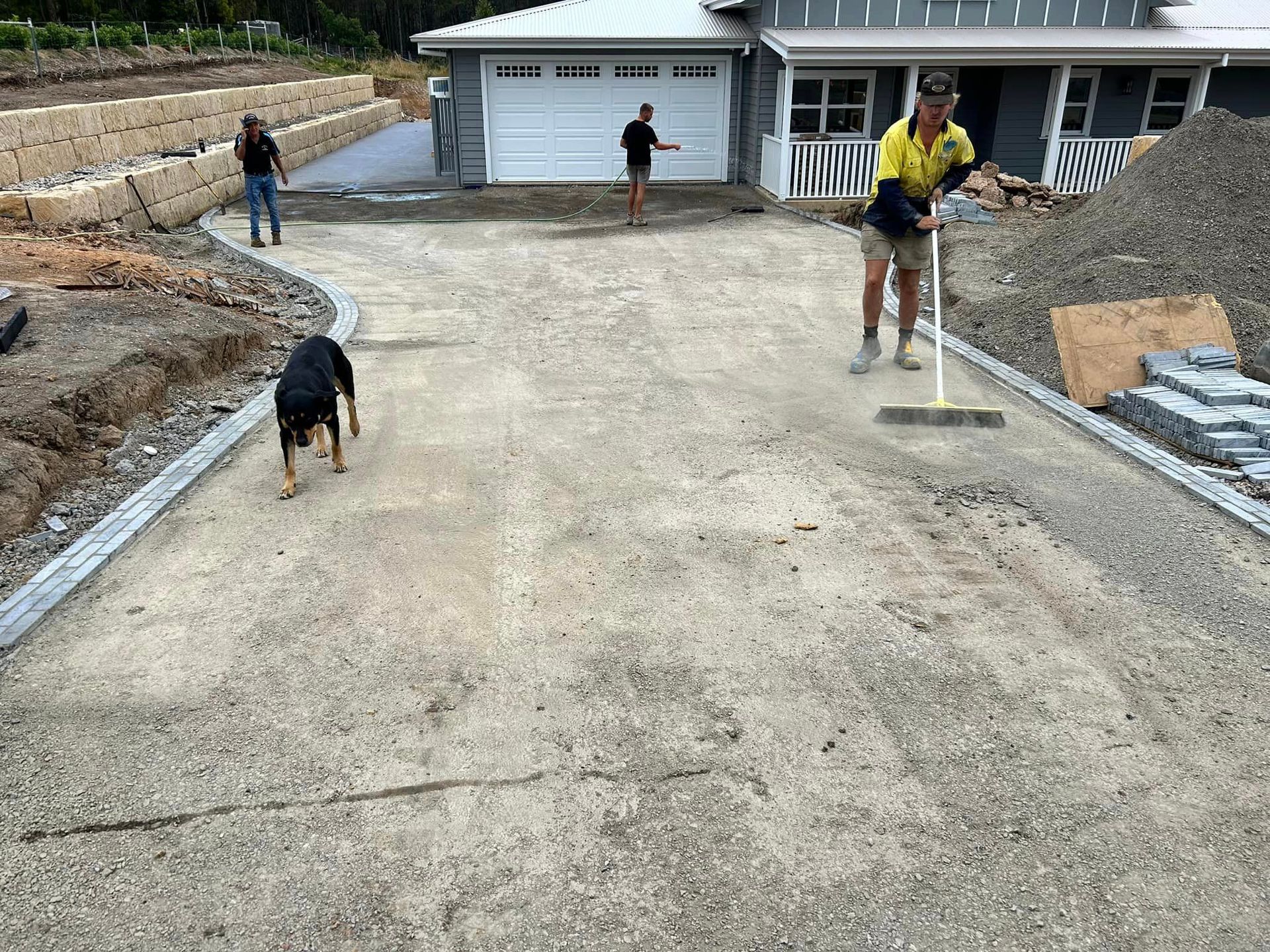 A man is sweeping a driveway in front of a house while a dog watches.