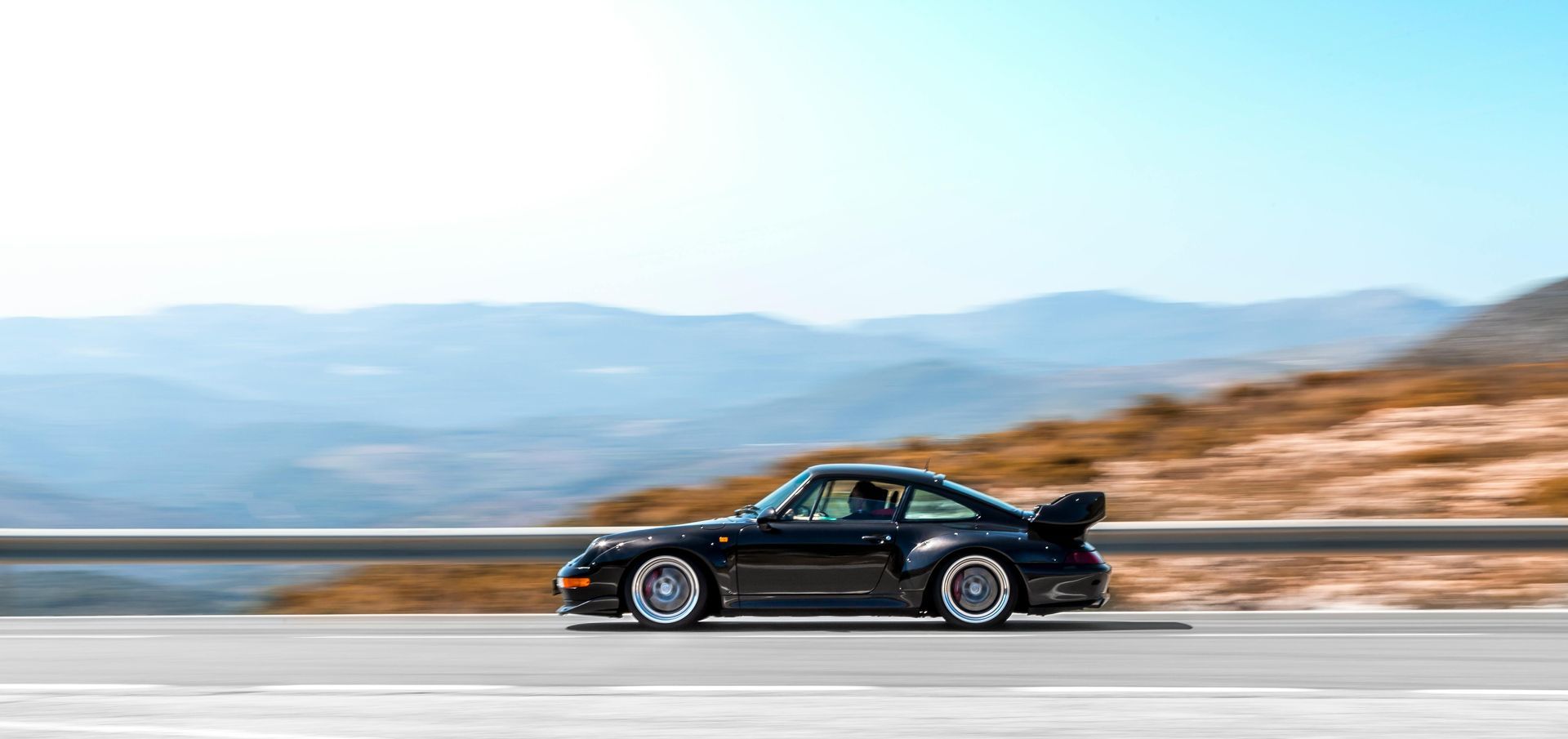 A black porsche is driving down a road with mountains in the background.