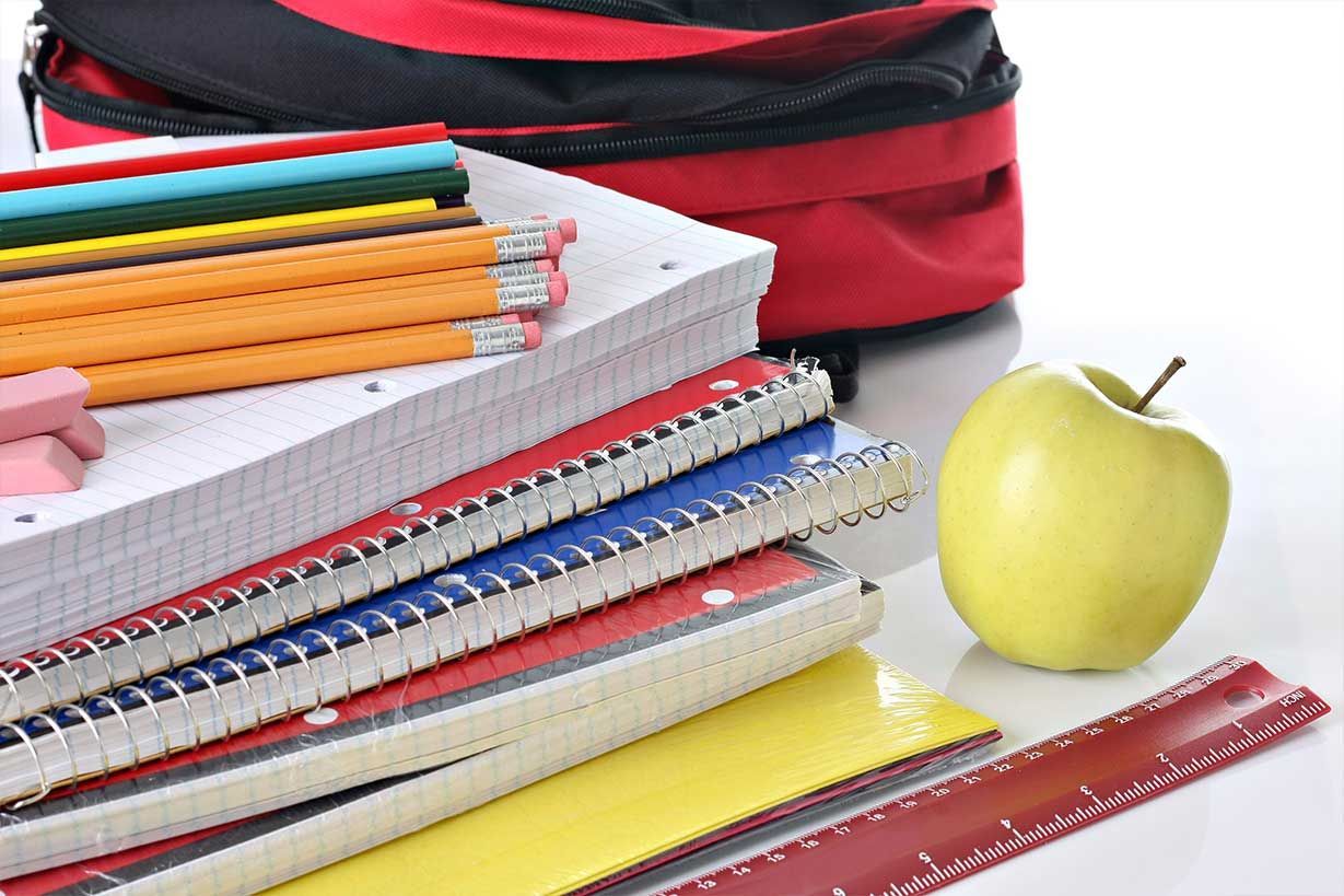 Una pila de cuadernos, lápices y una regla junto a una manzana y una mochila.