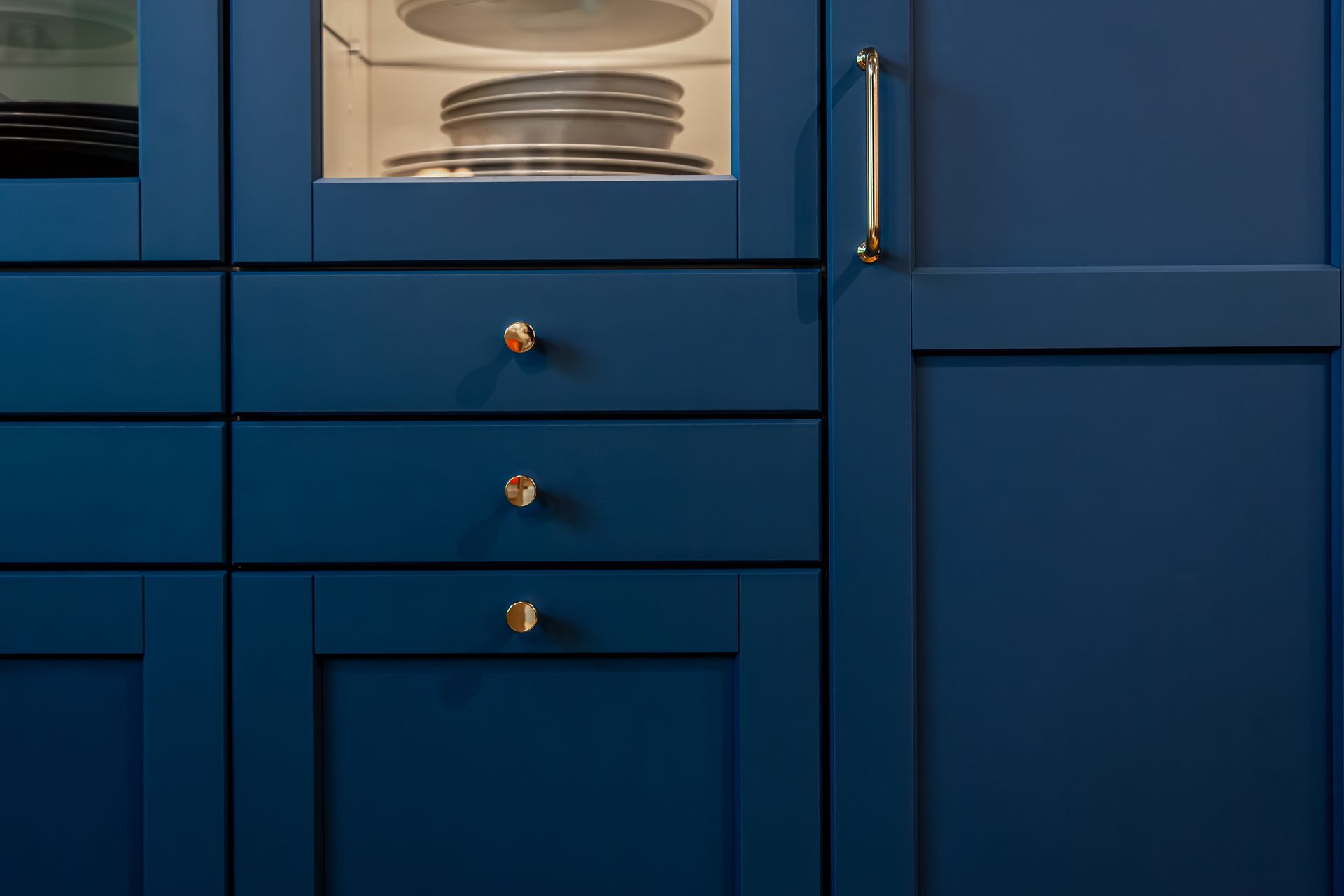 Dark blue kitchen cabinetry featuring a glass-front display cabinet with stacked plates and brass hardware.