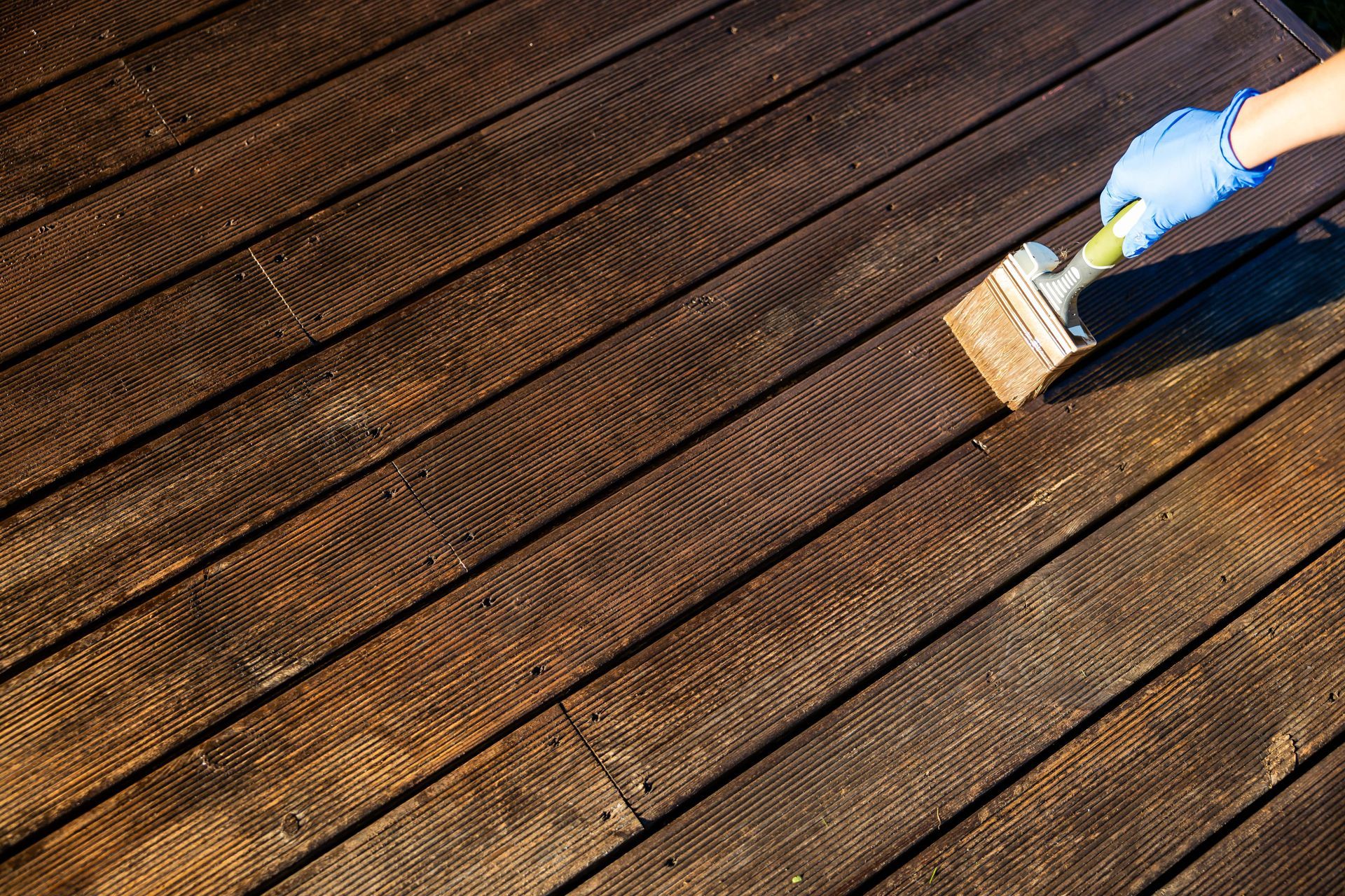 A hand wearing a blue protective glove uses a brush to apply wood stain to dark, textured wooden deck planks.